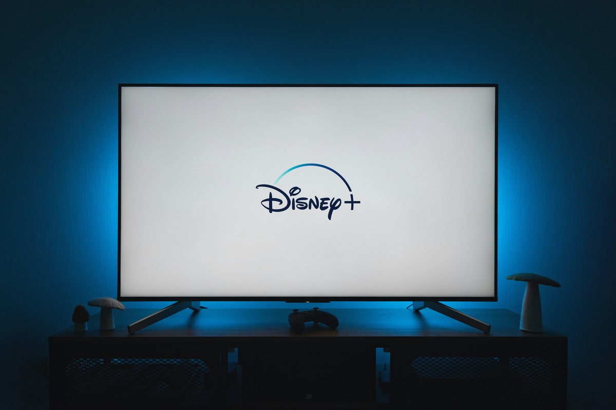 A wall-mounted television in a dark room showing the Disney+ logo, with a games controller on the media console below