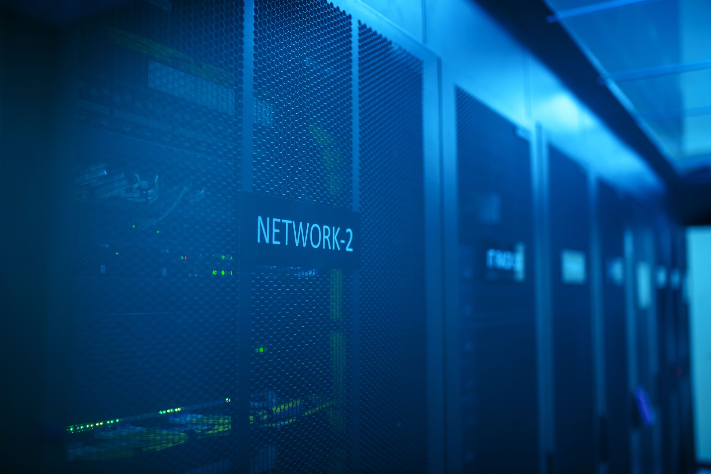 A dim, blue-lit datacentre aisle of server racks, one bay labelled NETWORK-2 — the kind of room a rented game server actually sits in, and the choice of which room decides everyone's ping