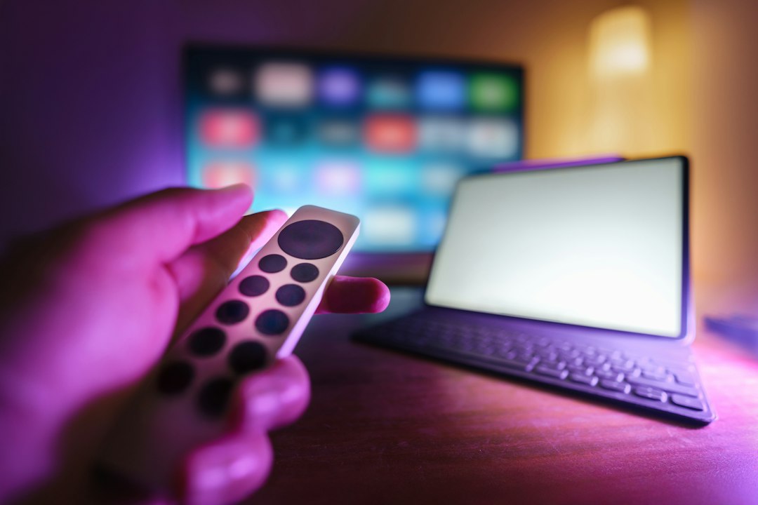 A hand pointing a streaming remote at a TV showing a grid of streaming apps, with a tablet on the desk beside it