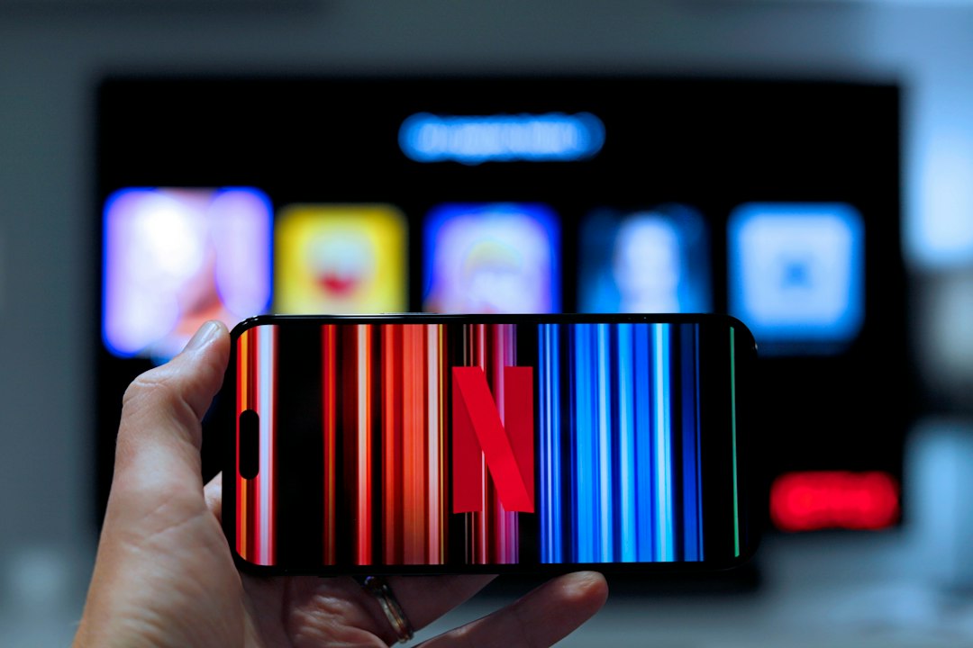 A hand holding a phone showing the Netflix logo in front of a blurred TV screen of streaming app icons
