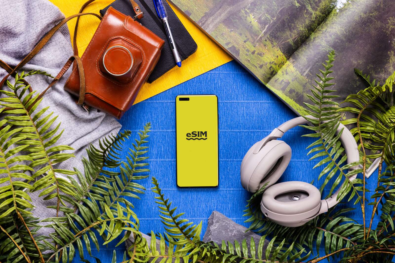 Travel flat lay with a smartphone showing an eSIM screen, headphones, camera and a map among ferns